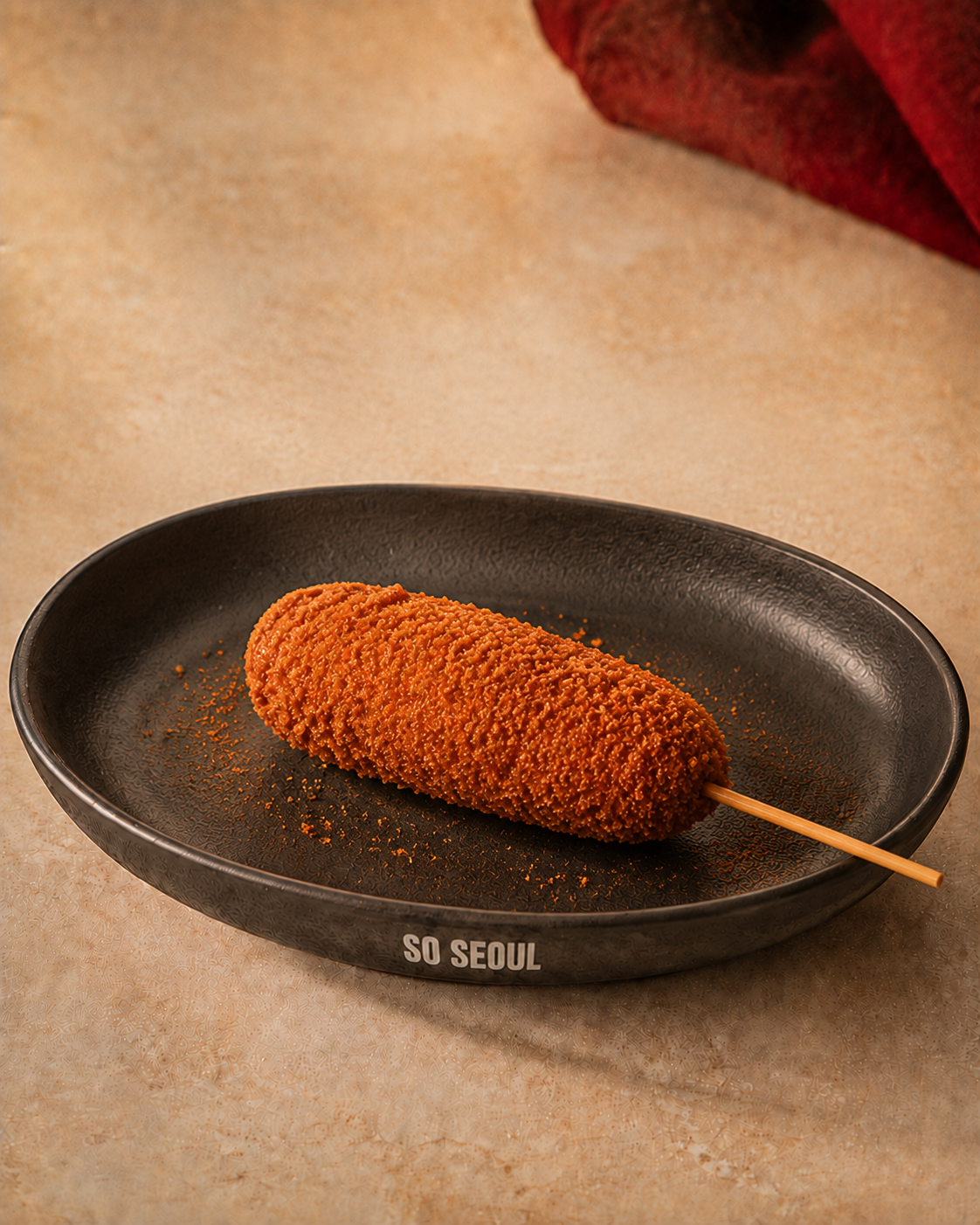 A single So Seoul Korean corn dog on a dark plate with scattered peri-peri seasoning.