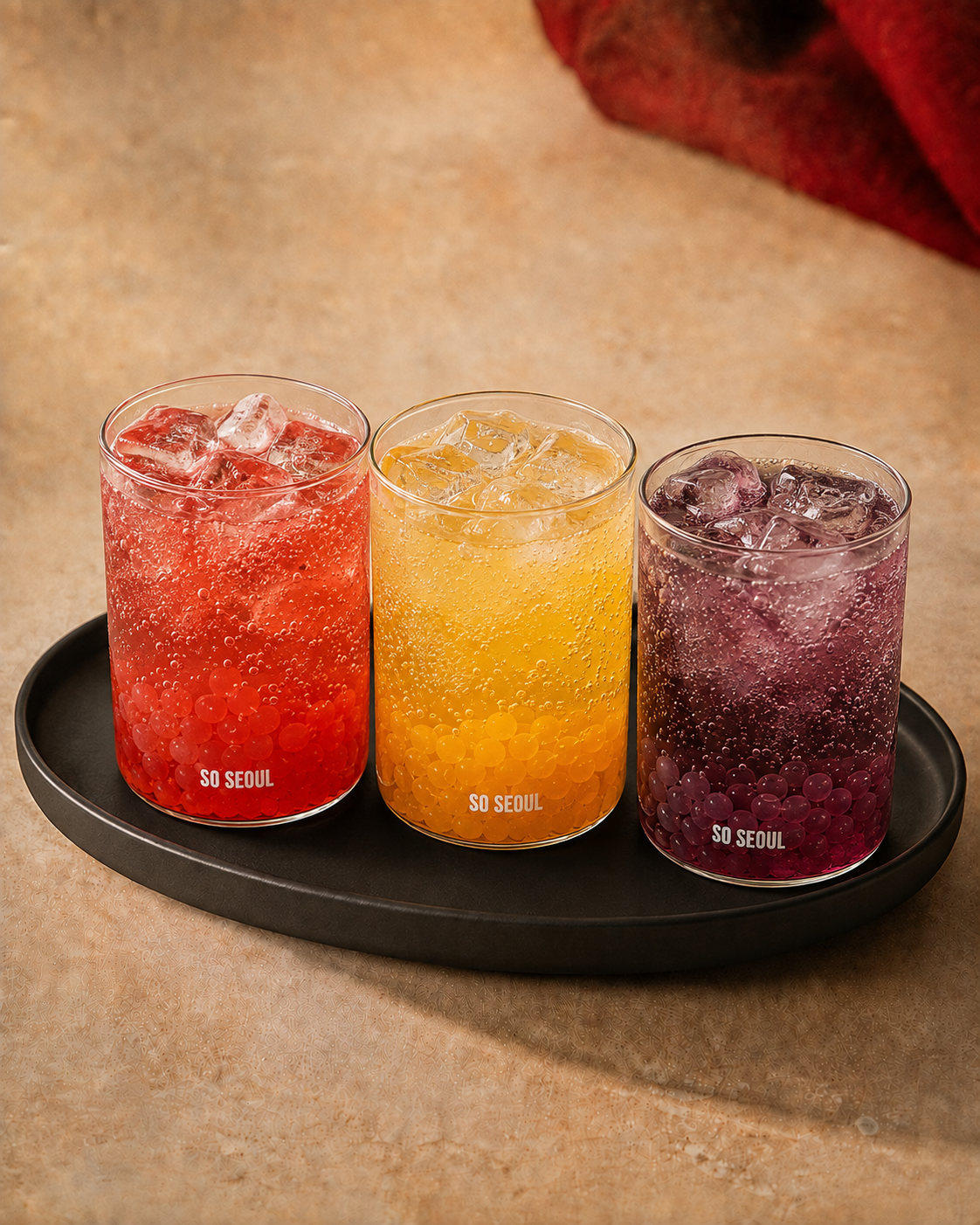 Three So Seoul popping boba drinks over ice — strawberry, mango and blackcurrant — on a black tray.